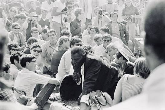 A historic black and white photo depicting a large crowd gathered outdoors, with people sitting on the grass and others standing around. In the foreground, a man in a suit is speaking to the audience, while numerous attendees appear engaged and attentive. The scene conveys a sense of community and participation during an event.