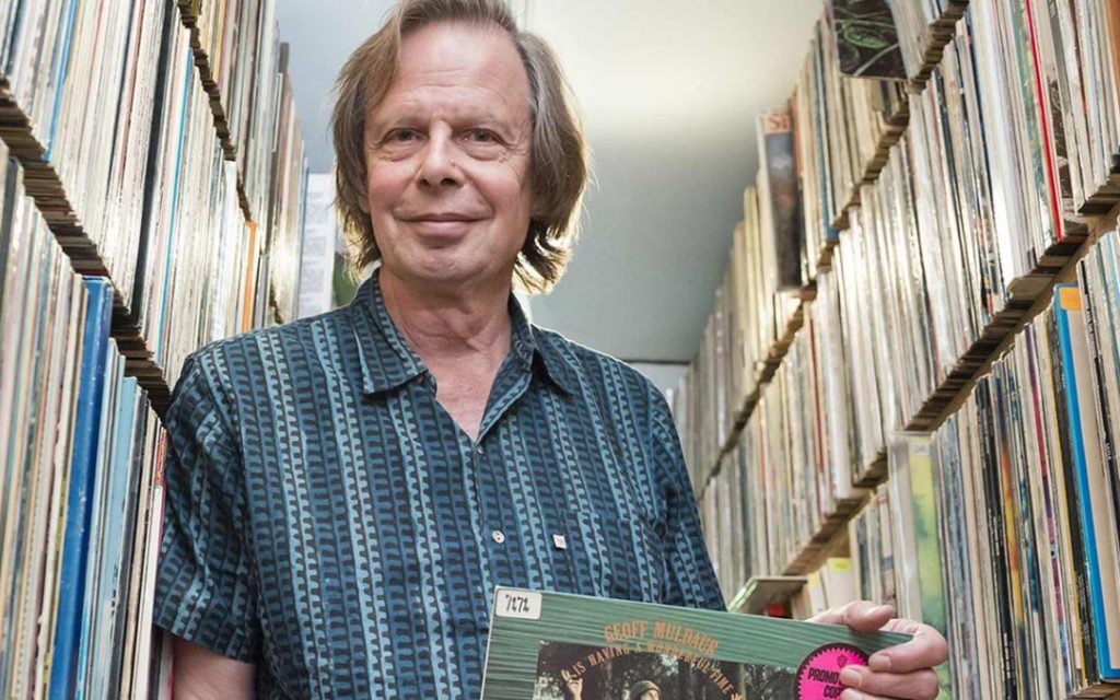 A man holding a vinyl record in a narrow aisle surrounded by stacks of vinyl albums.